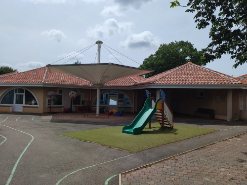 Cour de r&eacute;cr&eacute;ation des petits avec petit toboggan vert et escalier color&eacute; sur sol souple. B&acirc;timent scolaire beige &agrave; toiture rouge avec voile d'ombrage au centre de la cour.