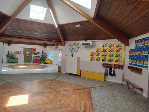 Hall d'entr&eacute;e de l'&eacute;cole maternelle avec charpente bois apparente et puits de lumi&egrave;re. Sols parquet et carrelage, murs d&eacute;cor&eacute;s de dessins d'enfants color&eacute;s, vestiaires avec pat&egrave;res le long du mur.